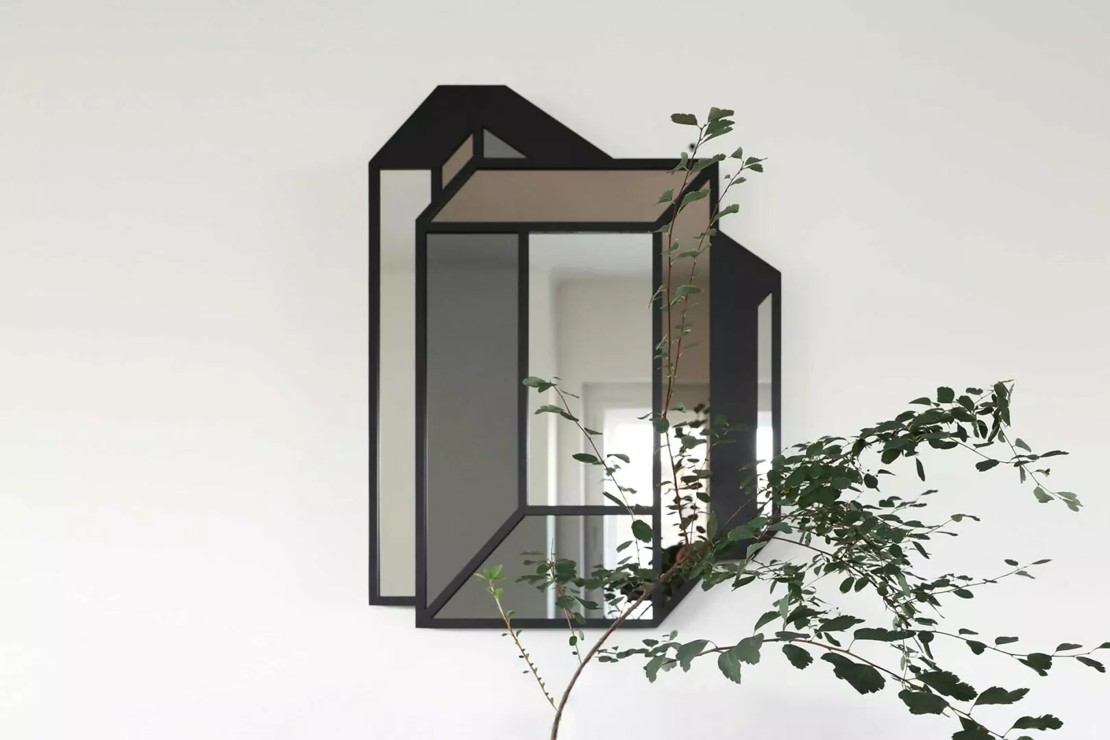 Modern geometric mirror with green plant on white wall.